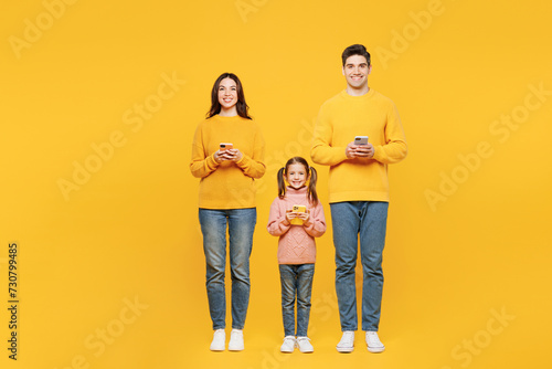 Full body young happy fun parents mom dad with child kid girl 7-8 years old wear pink sweater casual clothes hold in hand use mobile cell phone isolated on plain yellow background. Family day concept. photo