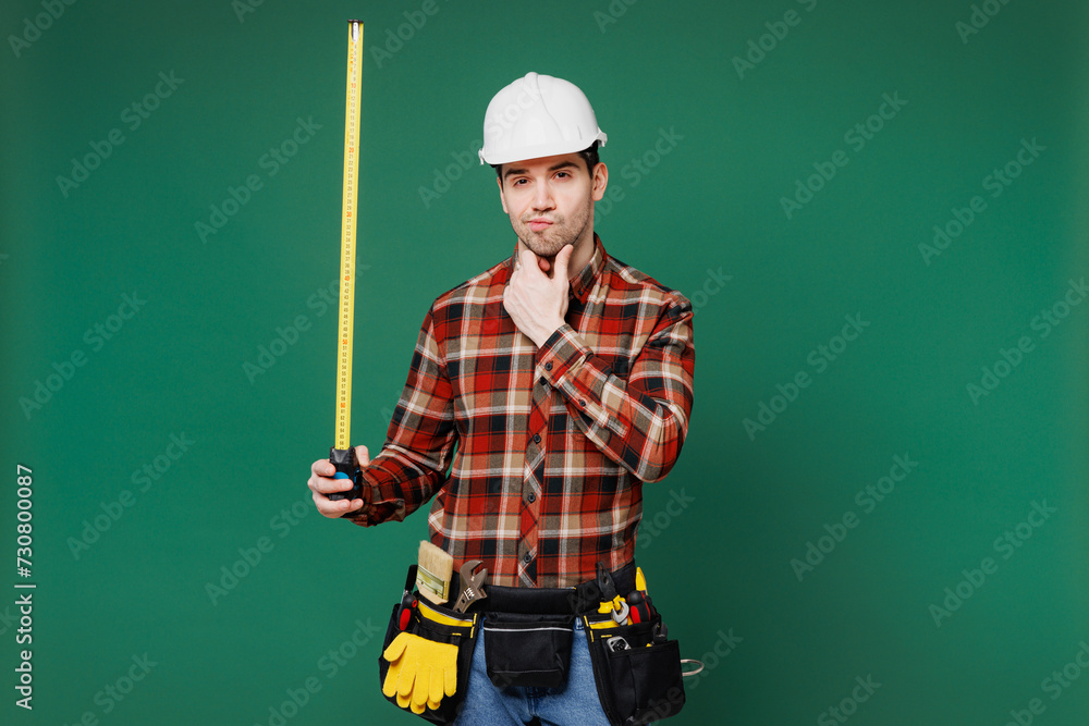 Young minded employee laborer handyman man wear red shirt hardhat hat hold tape measure isolated on plain green background. Instruments accessories for renovation apartment room. Repair home concept. Young minded employee laborer handyman man wear red shirt hardhat hat hold tape measure isolated on plain green background. Instruments accessories for renovation apartment room. Repair home concept.