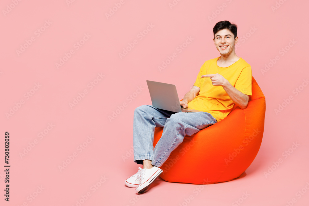 Full body young IT man wearing yellow t-shirt casual clothes sit in bag chair hold use work point finger on laptop pc computer isolated on plain pastel light pink background studio. Lifestyle concept.