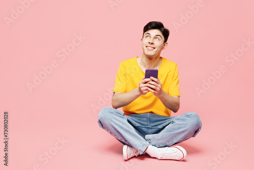Full body happy young man he wearing yellow t-shirt casual clothes sits hold in hand use mobile cell phone look aside on area isolated on plain pastel light pink background studio. Lifestyle concept. © ViDi Studio