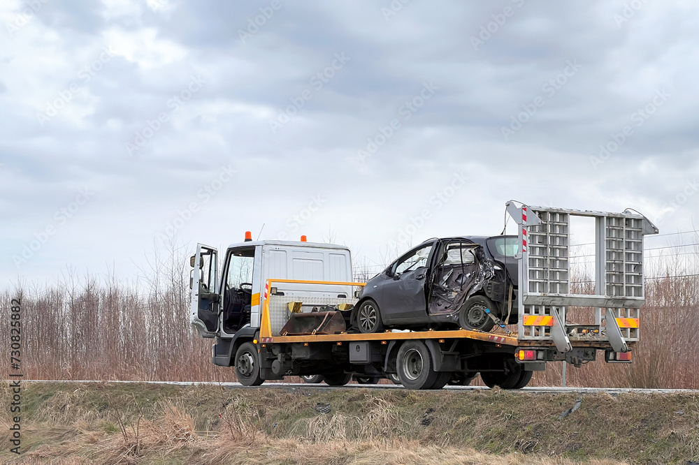 A side-impact collision leaves a car in need of a tow truck.  The car is severely damaged and may be beyond repair. A modern black Japanese hatchback is being hauled away by a tow truck on the highway