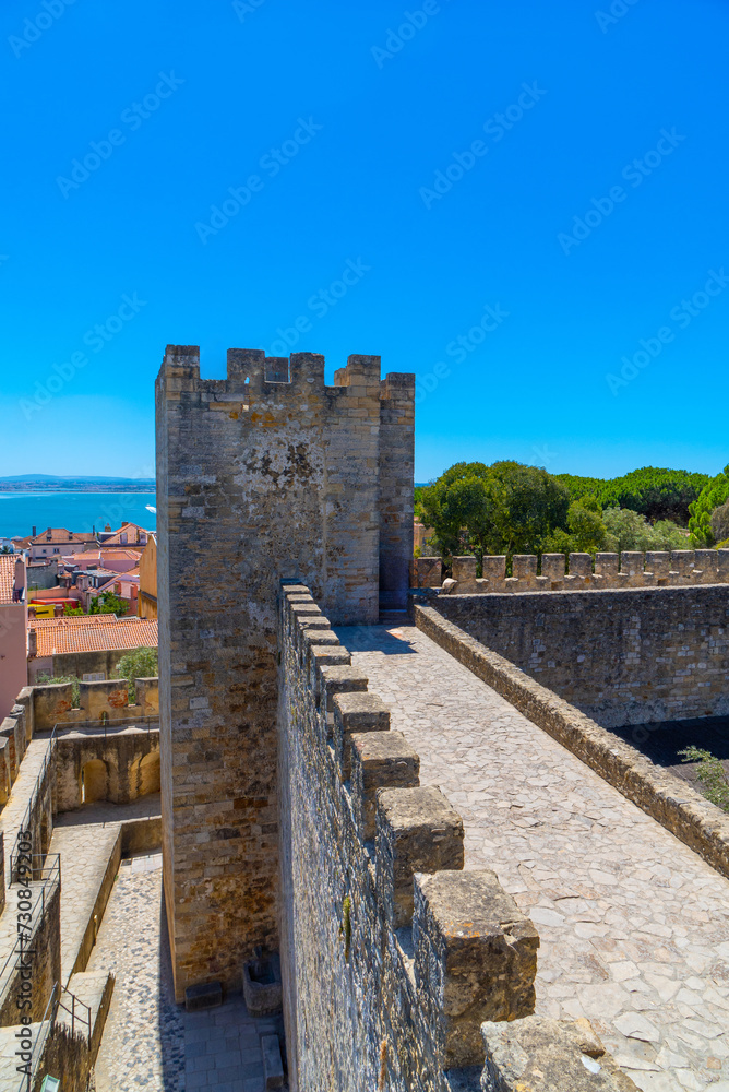 Top view of the walls, barbican, wall and walkway to the watchtower or tribute of the Castle of Saint George with part of the typical houses of Lisbon, and the Tagus River in the background.