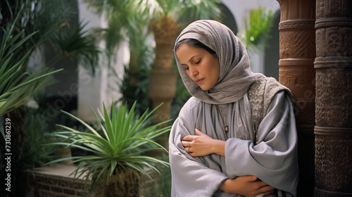 Portrait of a serene woman in a gray jellaba, hands softly cradling her face, eyes closed with a content smile, standing before a peaceful Riad courtyard garden photo