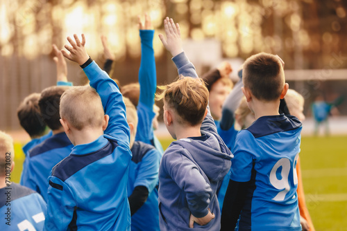 Children's sports team standing in a circle and rising hands up. Kids play sports match. School boys in blue jersey shirts huddling in a team. School sports team compete in a game photo