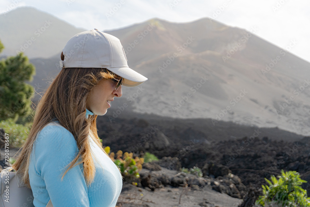woman with sunglasses and cap looks at the ground in front of volcano