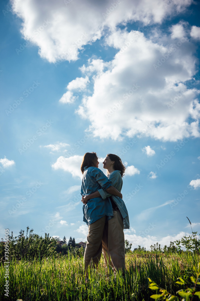 Two beautiful lesbian girlfriends in blue shirts are standing and hugging on the grass against a cloudy blue sky. The concept of love and happiness of LGBT people Two beautiful lesbian girlfriends in blue shirts are standing and hugging on the grass against a cloudy blue sky. The concept of love and happiness of LGBT people