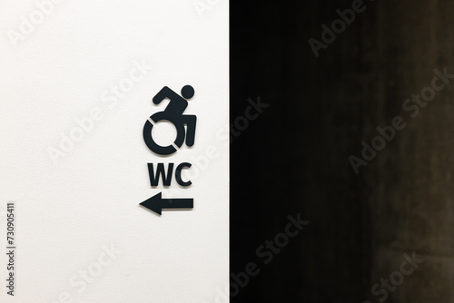 A minimal black restroom sign with an arrow for persons with disabilities on a flat white wall with concrete wall on the background