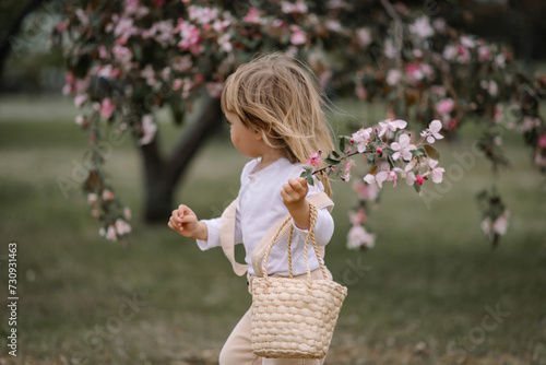 A little girl, a child in beige pants and a white blouse, with a basket on her shoulder, runs through the green grass with a twig in her hand. Pink blossoming apple tree on a blurred background