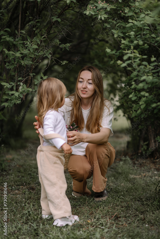 Fototapeta premium A small stylish girl, a child and her mother sat down on a green lawn in a park among bushes. Mom hugs her daughter. The daughter is holding a basket and a branch of a blossoming apple tree.
