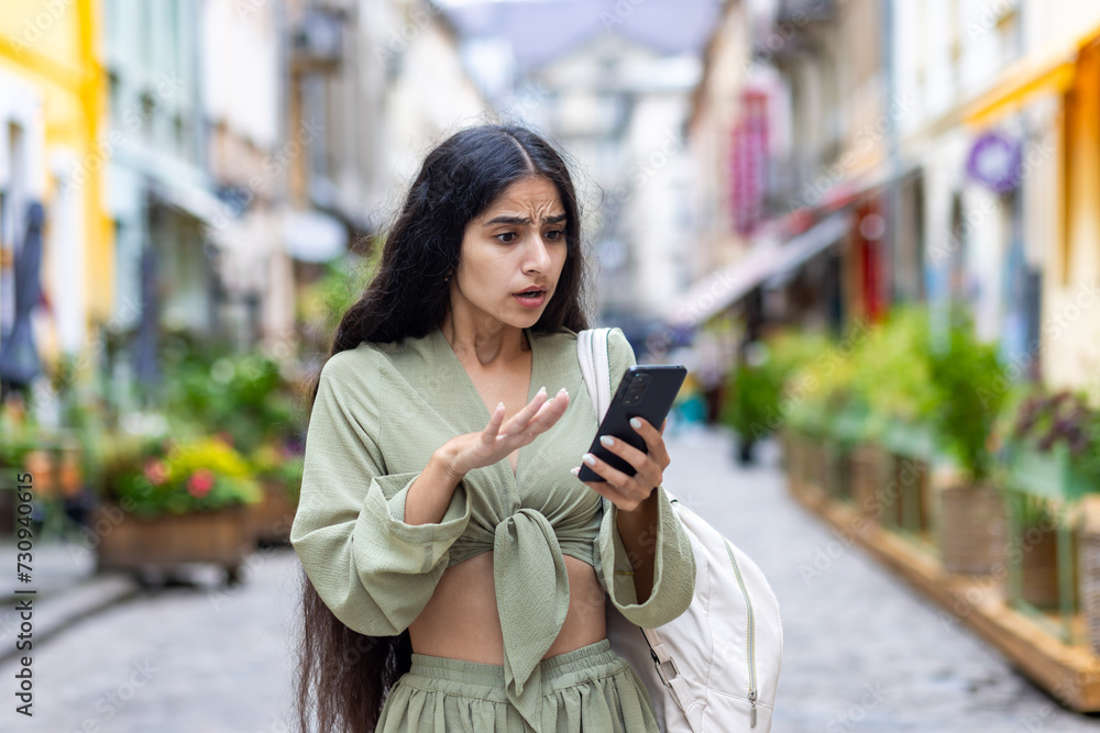 Shocked and disappointed young Indian student girl standing on city street and looking at mobile phone screen pointing with hand