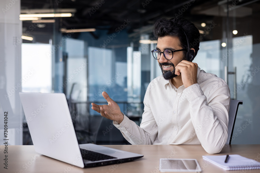 A smiling Muslim man works in the office in a headset and on a laptop, holds an online meeting with clients and partners, discusses issues and a project A smiling Muslim man works in the office in a headset and on a laptop, holds an online meeting with clients and partners, discusses issues and a project