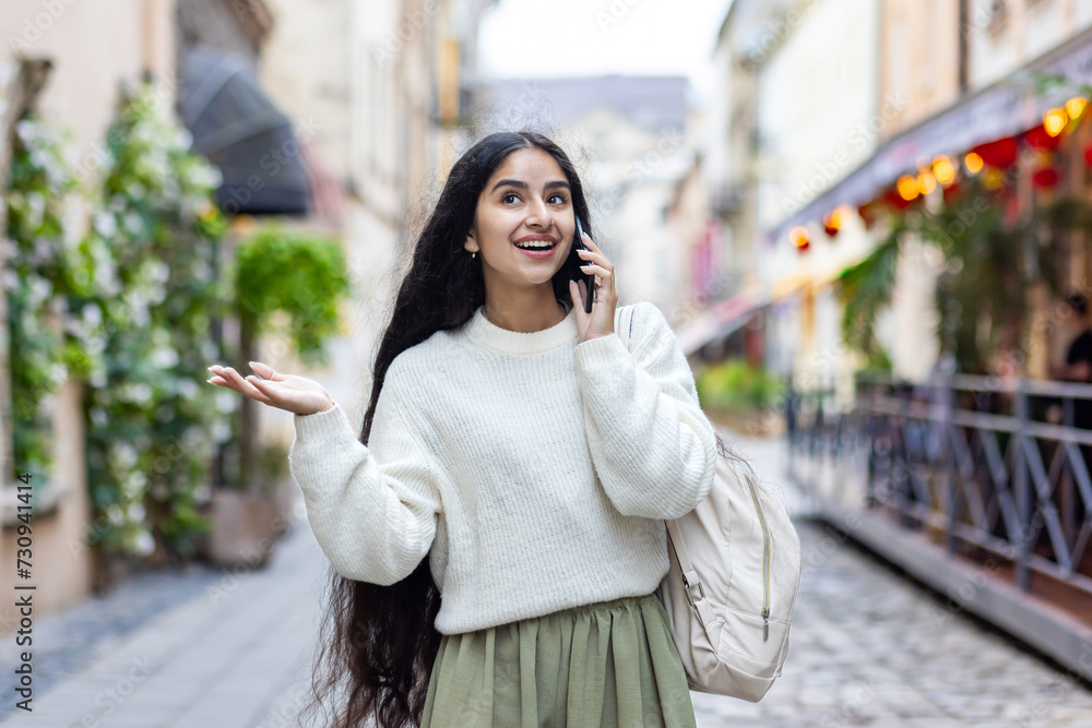 Fototapeta premium An Indian young smiling woman walks around the city, talks expressively on a mobile phone, has an emotional discussion, gestures with her hands