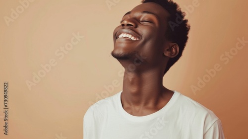 A young man with a joyful expression smiling broadly with his eyes closed wearing a white t-shirt against a warm soft-focus background.