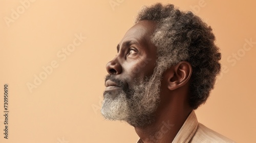 A contemplative man with a gray beard and curly hair looking upwards with a thoughtful expression set against a soft warm-toned background.