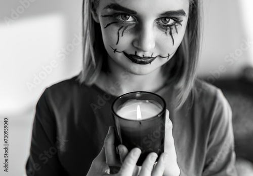 Preteen girl with spooky Halloween makeup posing at camera with lighting candle. Kid's portrait for creepy holiday in black and white.