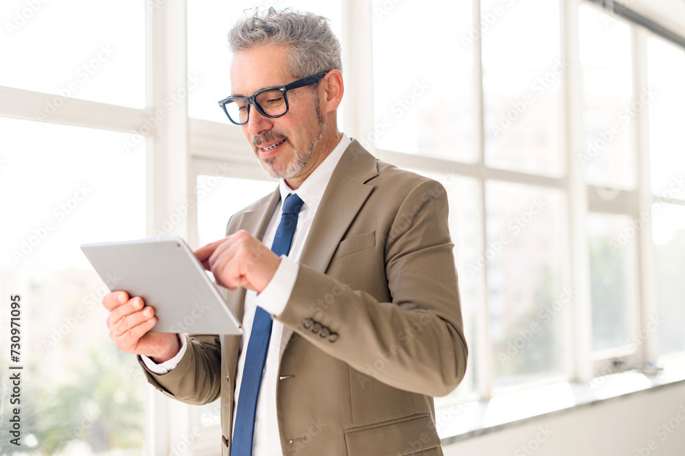 The seasoned businessman is interacting with digital tablet in his office, a symbol of his adaptability to the evolving business landscape. Mature grey-haired man using tablet for online comunication