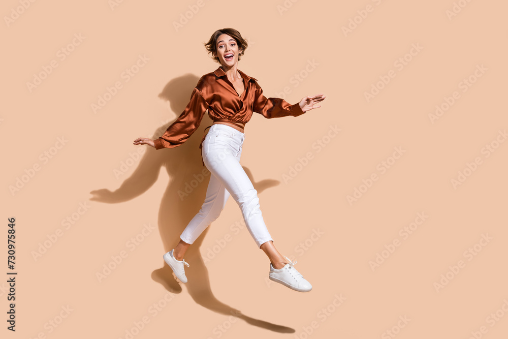 Full length photo of excited pretty woman wear brown blouse jumping high walking empty space isolated beige color background