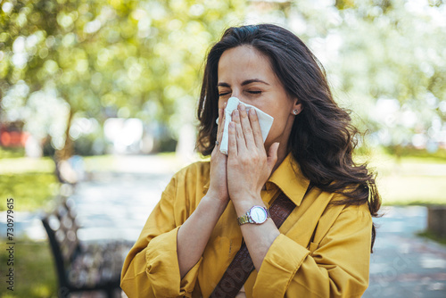 Shot of an attractive young woman feeling ill and blowing her nose with a tissue outdoors. Woman has sneezing. Young woman is having flu and she is sneezing. Sickness, seasonal virus problem concept photo