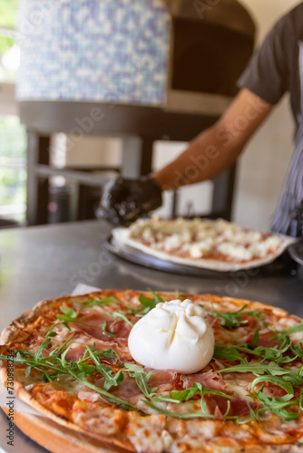 Burrata cheese topping on Italian pizza with prosciutto and arugula, served on the wooden board with traditional oven on background.