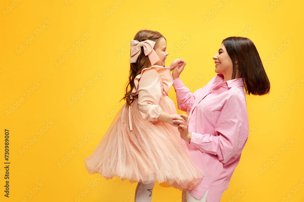 Birthday celebration. Stylish happy mother dancing with her little pretty daughter, kid against yellow studio background. Concept of happiness, Mother's day, childhood, fashion and lifestyle Birthday celebration. Stylish happy mother dancing with her little pretty daughter, kid against yellow studio background. Concept of happiness, Mother's day, childhood, fashion and lifestyle