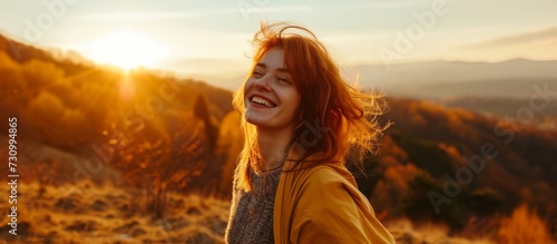 A happy woman with a smile stands on a grassy hill at sunset, enjoying the beautiful landscape of the ecoregion under the colorful sky. © AkuAku