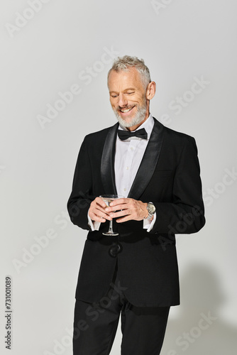 cheerful handsome mature man in chic tuxedo with gray beard smiling and holding martini with olives