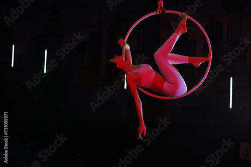 Young woman performing acrobatic element on aerial ring against dark background. Space for text photo