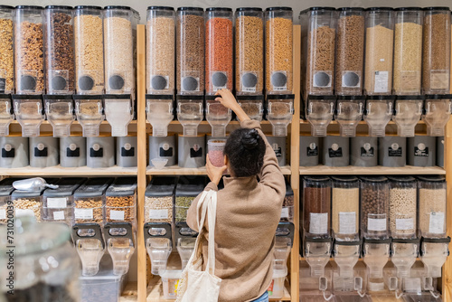 African American woman refilling reusable package with bulk products in local zero waste grocery store. Rear view of woman buying grains and cereals in sustainable eco grocery store.