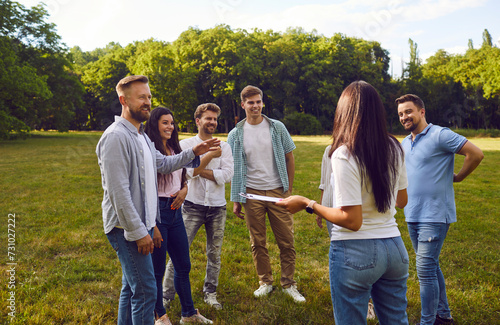 Group of happy friends or corporate coworkers standing on grass in summer park on weekend and listening to manager with clipboard listing their tasks and explaining rules of outdoor team building game photo