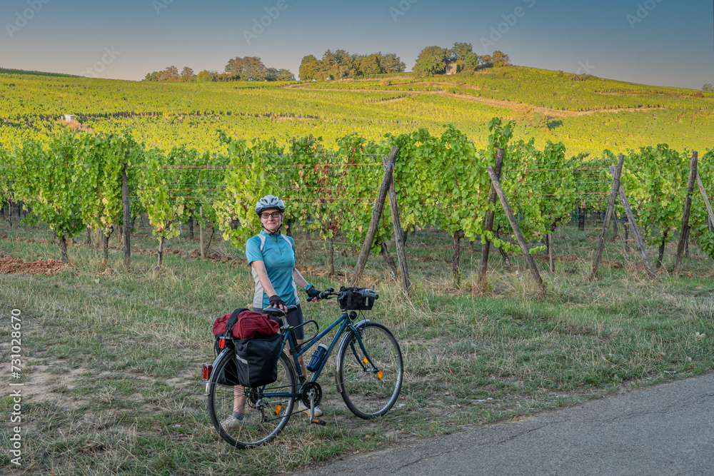 Fototapeta premium Mittelbergheim, France - 09 10 2020: Alsatian Vineyard. A woman dressed in a blue t-shirt, black shorts and a helmet, poses next to her blue bicycle with vine fields behind.