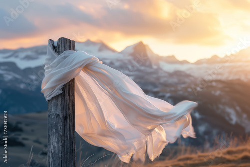 A white silk scarf fluttering in the wind on a wooden cross overlooking a valley between snow-capped mountain peaks at sunset. photo