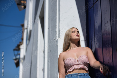 Young woman, blonde, green eyes, wearing pink top and jeans, looking at the sky, leaning against the frame of a white Andalusian house, receiving the sun's rays. Concept beauty, peace, sunny.