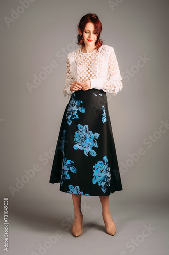 Long sleeve blouse in white and midi skater skirt in black. Studio portrait of shy young woman on high heels. Female evening outfit. Ginger lady smiling on studio background.