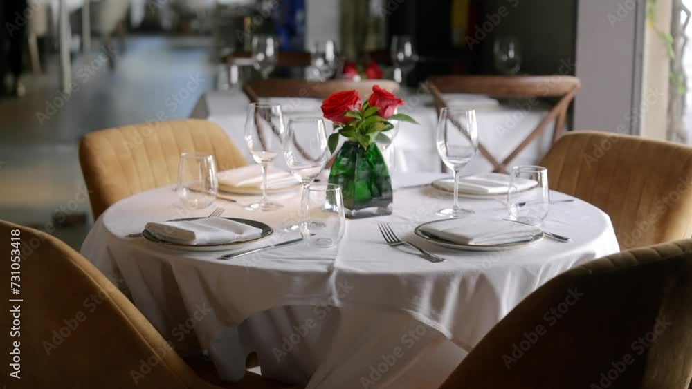 Well-set round table covered with white tablecloth in restaurant. Each place including plate, nappkin and two glasses. Bouquet of red roses in green vase Well-set round table covered with white tablecloth in restaurant. Each place including plate, nappkin and two glasses. Bouquet of red roses in green vase