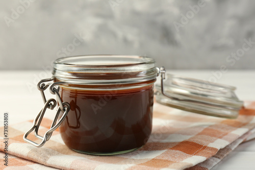 Tasty barbecue sauce in glass jar on white wooden table, closeup