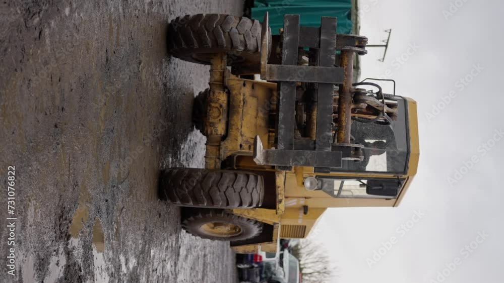 A yellow forklift, a machine used in mechanical engineering, with composite material and car tires, is parked by the road, next to a container. Vertical video A yellow forklift, a machine used in mechanical engineering, with composite material and car tires, is parked by the road, next to a container. Vertical video