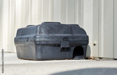 Rat bait station by a wall outdoors, front view at ground level. Common pest control solution for commercial properties.