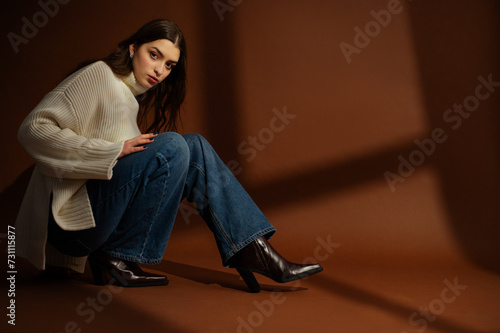 Fashionable confident woman wearing oversized white sweater, wide leg jeans, leather cowboy boots, posing on brown background. Full-length studio fashion portrait. Copy, empty blank space for text 