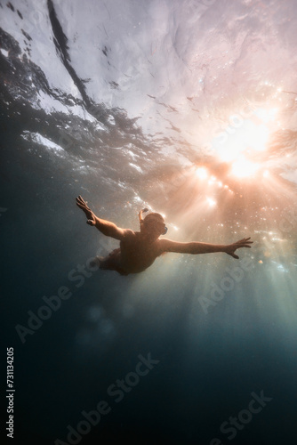 Person swimming underwater with sun rays shining through the surface.