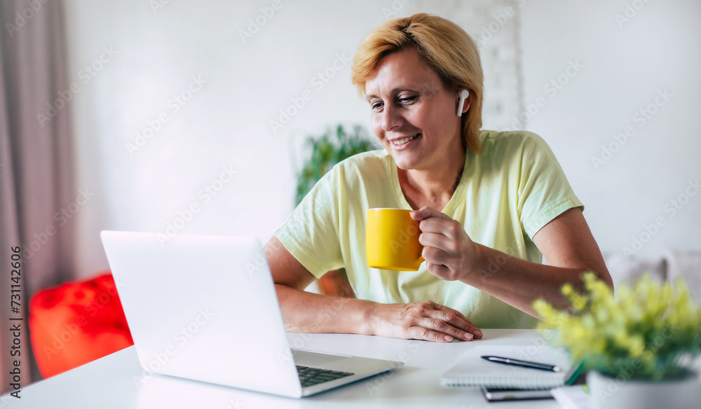 Portrait of happy middle-aged female with air Bluetooth headphones and beverage in hand is watching some movie on laptop at home. Online chat with family or friend Portrait of happy middle-aged female with air Bluetooth headphones and beverage in hand is watching some movie on laptop at home. Online chat with family or friend