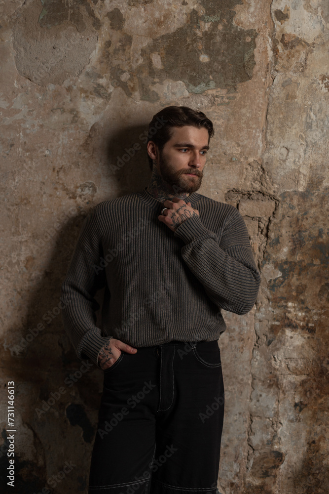 Handsome fashion hipster man with a beard in a fashionable knitted vintage sweater stands near a grunge texture wall