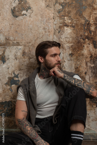 beautiful brutal hipster man with a beard and hairstyle with a tattoo in a white T-shirt with a sweater and jeans sits on the floor near a concrete grunge texture wall