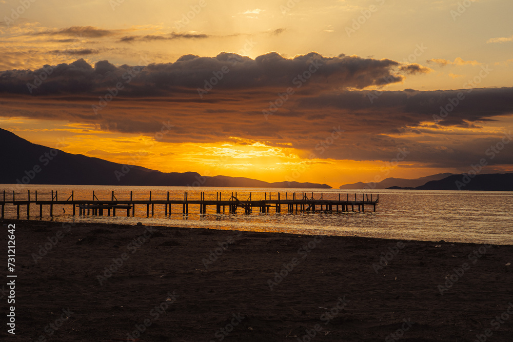 A dramatic sunset in the Mediterranean. The sun behind the clouds, beach and a dock in the foreground. 