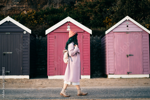 Stylish hipster woman with color hair in pink outfit and backpack walking along wooden beach huts on seaside. Off season Travel concept. Seasonal street fashion. Barbiecore style. Simple pleasures photo