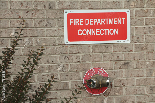 fire department connection sign on beige brick wall with two twin connections below from one spout in red circle auto spkr, white on red