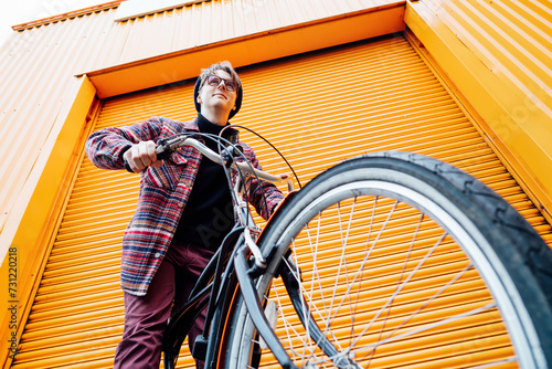 Bottom view Young stylish hipster man standing with a cruiser city Bicycle near orange Wall. Student Resting after ride bicycle. Eco friendly transportation for carbon footprint. Sustainable lifestyle