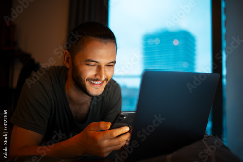 Young man lying on bed, using smartphone and laptop, evening time, dark photo.