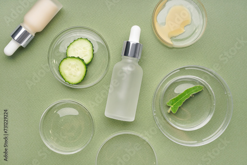 Petri dishes with sample, cucumber pieces and bottles of cosmetic products on green background