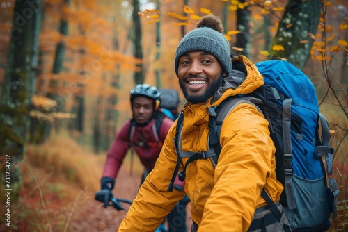 A beaming man, bundled in a cozy winter jacket and sporting a backpack and hat, stands tall with his trusty bicycle among the autumnal trees, ready for an outdoor adventure
