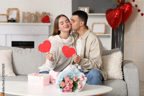 Beautiful young couple with gift box, bouquet of flowers and paper hearts celebrating Valentine's Day at home